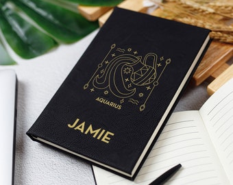 May include: Black leather journal with gold lettering. The front cover features a gold line drawing of the Aquarius zodiac sign with the word "Aquarius" and the name "JAMIE".
