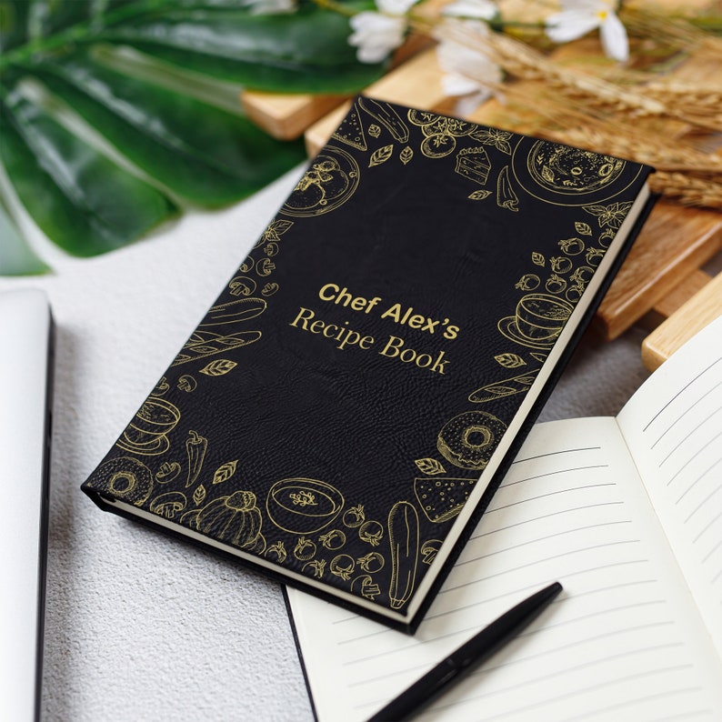 May include: Black recipe book with gold text and illustrations. The cover reads "Chef Alex's Recipe Book" and features drawings of food items. The book is open to a blank page next to a pen.
