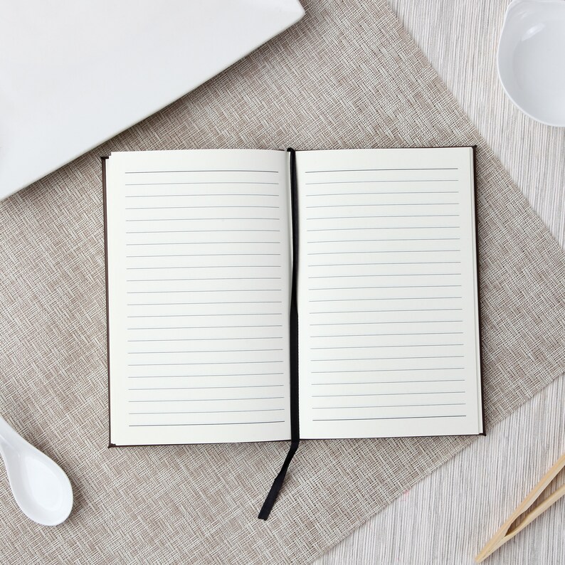May include: An open, lined notebook with a black ribbon bookmark. The notebook has a dark brown cover and is positioned on a textured, beige placemat. A white plate, bowl, spoon, and chopsticks are arranged around the notebook.
