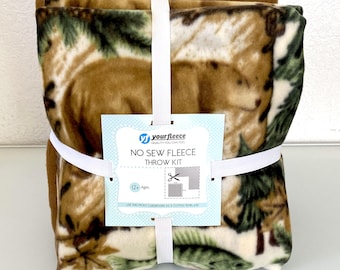 May include: A bundled fleece throw kit featuring a bear and forest design in shades of brown, green, and cream. The kit includes a no-sew fleece throw with a white elastic band and a tag that reads "No Sew Fleece Throw Kit".