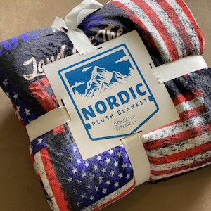 May include: A folded American flag patterned plush blanket with a white tag that says "Nordic Plush Blanket 50x60 in 127x152 cm".