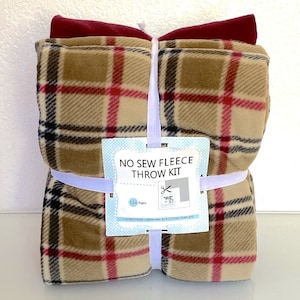 Peut inclure: Un kit de plaid en polaire sans couture, emballé et attaché avec un ruban blanc. La polaire présente un motif à carreaux beige, rouge et noir. Un morceau de polaire rouge foncé est sur le dessus. Le kit comprend une étiquette "NO SEW FLEECE THROW KIT".
