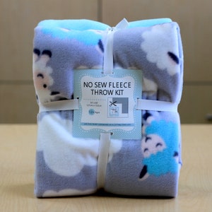 May include: A rolled-up fleece throw kit featuring a sheep pattern in shades of gray, white, and blue. The kit is tied with a white ribbon and elastic bands, and includes a tag that reads "NO SEW FLEECE THROW KIT". The throw measures 50 x 60 inches.