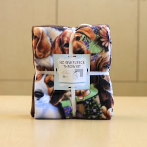 May include: A no-sew fleece throw kit featuring a dog print design. The kit includes two pieces of fleece fabric, a pattern, and instructions. The fabric is brown and white with a pattern of dogs. The kit is packaged and tied with white ribbon.