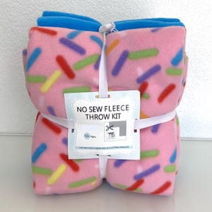 May include: A pink fleece throw kit with a colorful sprinkle pattern, tied with a white ribbon. The kit includes a blue border and a tag that reads "NO SEW FLEECE THROW KIT" and "12+ Ages".