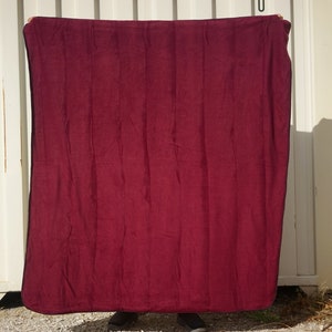 May include: A deep red fleece blanket with a black border. The blanket is being held up by a person's hands.