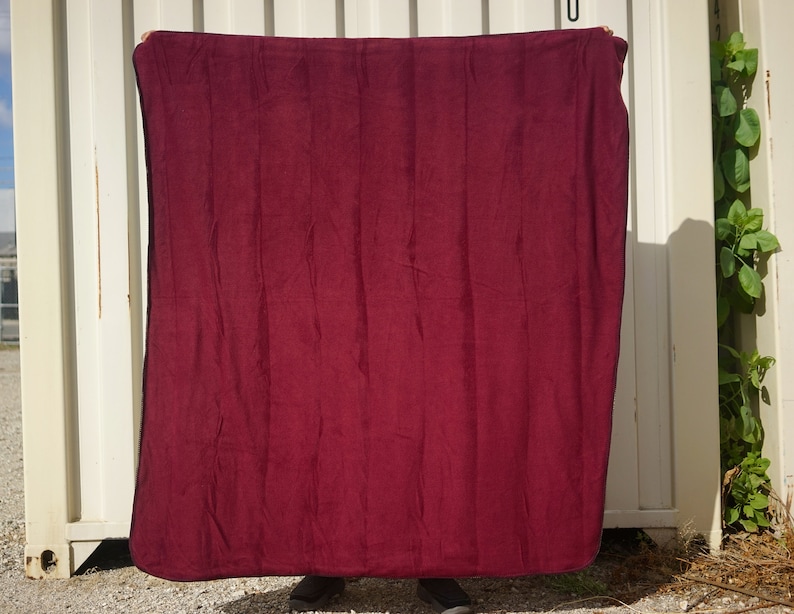 May include: A deep red fleece blanket with a black border. The blanket is being held up by a person's hands.