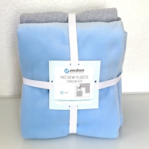 May include: A no-sew fleece throw kit, featuring a light blue and gray fleece fabric bundle, secured with a white elastic band. The kit includes a tag with the text "your fleece" and "NO SEW FLEECE THROW KIT".