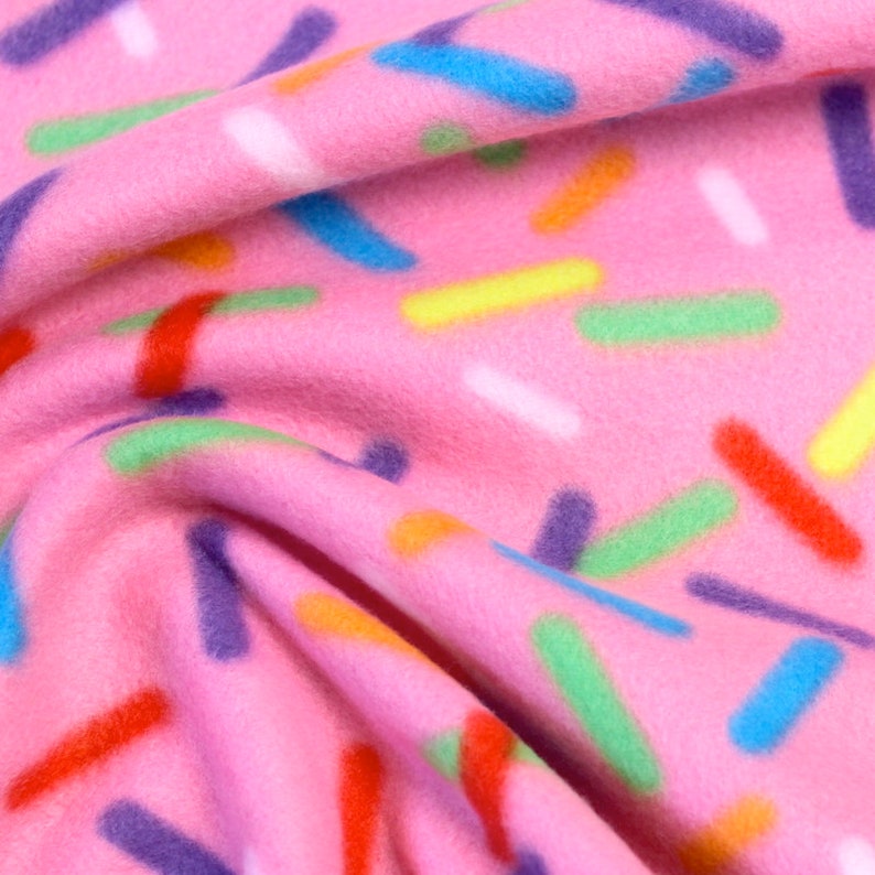 Puede incluir: Primer plano de tela polar rosa con un patr&oacute;n de chispas de colores. Las chispas son de varios colores, incluyendo azul, verde, amarillo, naranja, rojo y morado. La tela es suave y tiene una ligera textura.