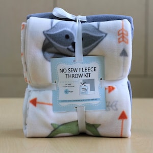May include: A no-sew fleece throw kit with a raccoon and arrow design. The kit includes two pieces of fleece fabric, a cardboard template, and instructions. The fabric is white with gray, orange, and green accents. The kit is 50 inches by 60 inches (127 cm x 152 cm) and is suitable for ages 12 and up.