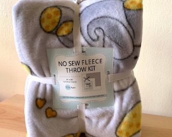 May include: A gray fleece throw kit, tied with a white ribbon. The blanket has a pattern of gray elephant shapes with yellow accents. The label reads "NO SEW FLEECE THROW KIT" and the blanket measures 50" x 60".