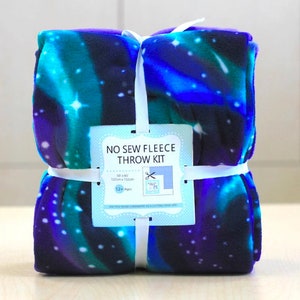 May include: A rolled-up no-sew fleece throw kit. The blanket features a vibrant blue, teal, and purple galaxy design with white stars. The kit includes a tag with the text "NO SEW FLEECE THROW KIT" and dimensions of 50 x 60 inches.