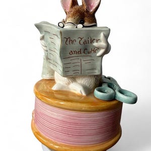 May include: A ceramic figurine of a brown and white mouse wearing glasses, reading a newspaper titled The Tailor and Cut, perched atop a large spool of pink thread with a pair of light blue ceramic scissors resting on the side of the spool base.