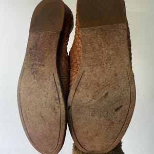 May include: A pair of worn, brown leather flats, soles facing up. The soles show wear and tear, with visible scuffs and discoloration. The shoes have a woven design on the upper part. The image is taken from a low angle, highlighting the soles and the shoe's construction.