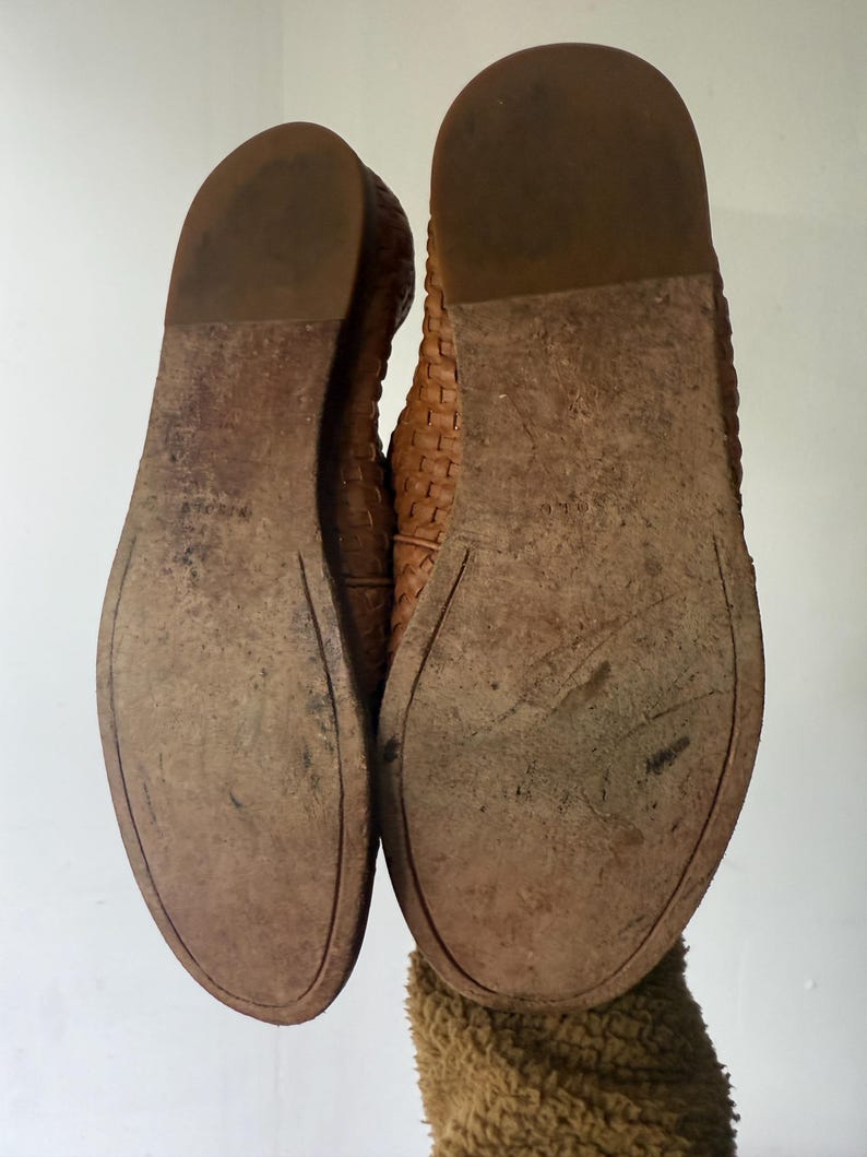 May include: A pair of worn, brown leather flats, soles facing up. The soles show wear and tear, with visible scuffs and discoloration. The shoes have a woven design on the upper part. The image is taken from a low angle, highlighting the soles and the shoe's construction.