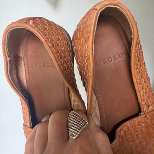 May include: A pair of woven leather loafers in a warm tan color. The interior of the shoe is a darker brown, with the brand name "ISOLO" visible. The shoes are held by a hand wearing a silver ring.