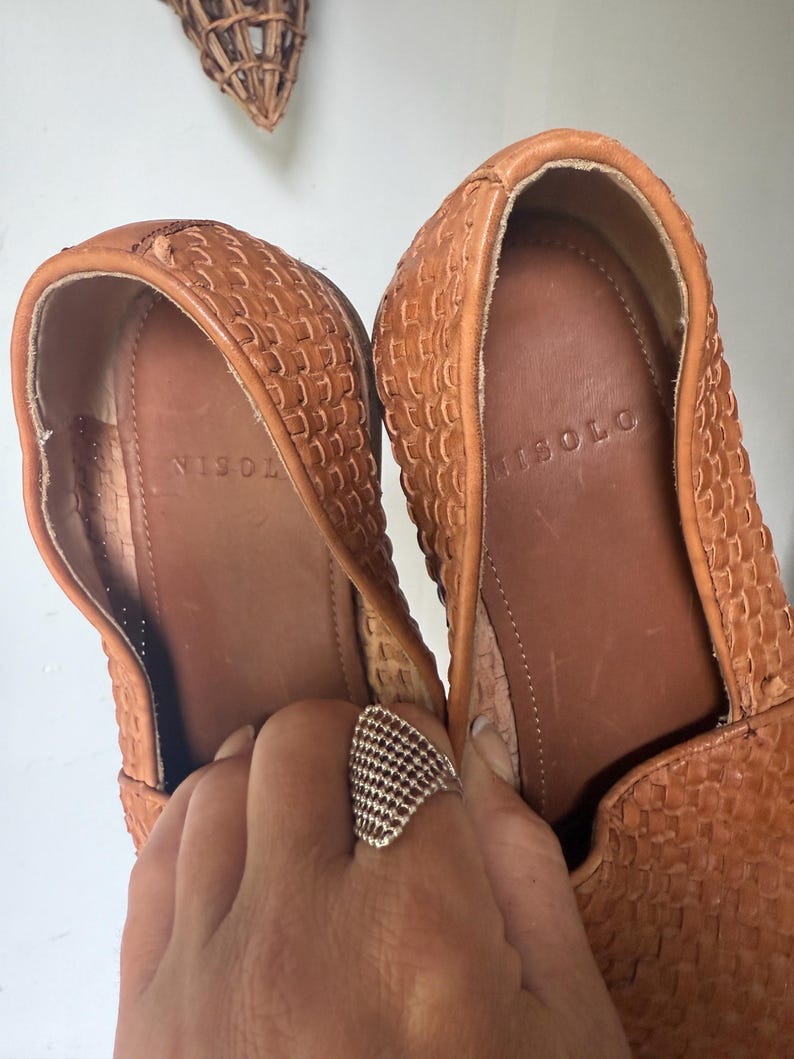 May include: A pair of woven leather loafers in a warm tan color. The interior of the shoe is a darker brown, with the brand name "ISOLO" visible. The shoes are held by a hand wearing a silver ring.