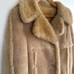 Puede incluir: Un abrigo de ante color beige con forro y ribete de piel de oveja. El abrigo tiene un cuello ancho, cierre de botones y bolsillos. Cuelga de una percha de madera contra una pared blanca.