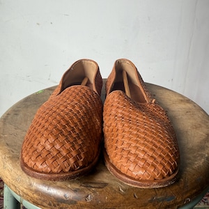 May include: A pair of woven leather slip-on shoes in a warm brown hue. The shoes feature a basket-weave design across the top and sides. The shoes are displayed on a wooden stool.