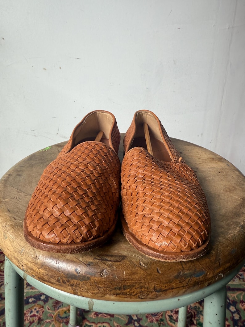 May include: A pair of woven leather slip-on shoes in a warm brown hue. The shoes feature a basket-weave design across the top and sides. The shoes are displayed on a wooden stool.