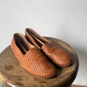 May include: A pair of woven leather loafers in a warm brown hue. The shoes have a slip-on design and a textured, basket-weave pattern. They are displayed on a wooden stool with a light green base.