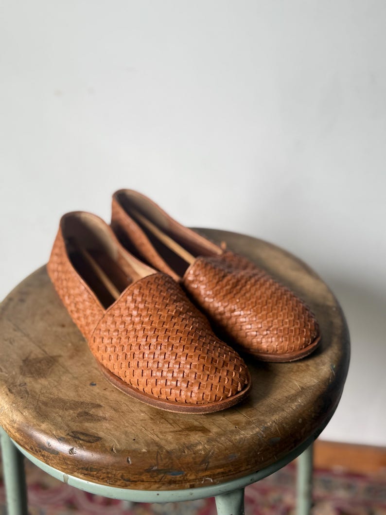 May include: A pair of woven leather loafers in a warm brown hue. The shoes have a slip-on design and a textured, basket-weave pattern. They are displayed on a wooden stool with a light green base.
