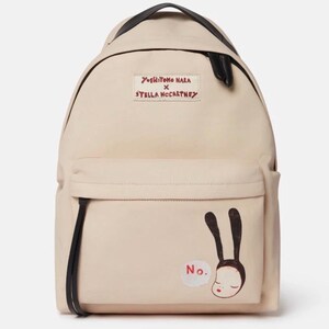 May include: A beige canvas backpack featuring a black top handle and zipper pulls. A rectangular patch reads Yoshitomo Nara x Stella McCartney. The front pocket displays a graphic of a character with long black ears and the red text No.