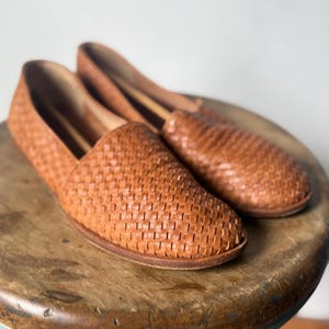 May include: A pair of woven leather loafers in a warm brown hue. The shoes have a classic slip-on design with a rounded toe and a textured, basket-weave pattern. They are placed on a wooden stool.