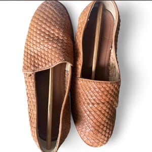 May include: A pair of brown woven leather loafers. The shoes have a slip-on design with a rounded toe and a woven pattern across the top. Shoe trees are inserted inside each shoe. The loafers are set against a white background.