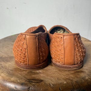 May include: A pair of brown woven leather loafers. The shoes have a basket weave design and a rounded toe. The soles are a darker brown. The shoes are sitting on a wooden stool.