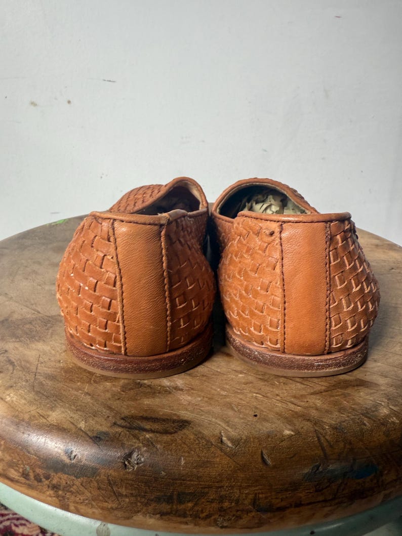 May include: A pair of brown woven leather loafers. The shoes have a basket weave design and a rounded toe. The soles are a darker brown. The shoes are sitting on a wooden stool.
