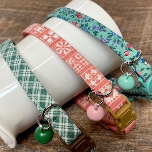 May include: Four cat collars with different patterns and colors. One is green plaid, another is coral with a floral design, and the third is teal with a floral pattern. Each collar has a matching bell and a plastic buckle.