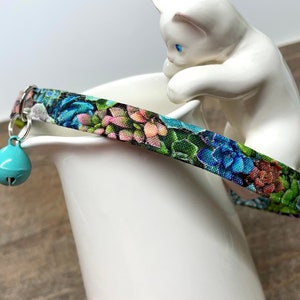 Może przedstawiać: Turkusowy obroża dla kota z dzwoneczkiem i kwiatowym wzorem przedstawiającym sukulenty. Obroża jest owinięta wokół białej ceramicznej figurki kota.