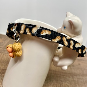 May include: A white ceramic cat figurine perched on a white mug, with a black and tan leopard print collar. A small peanut charm hangs from the collar. The mug is on a wooden surface.