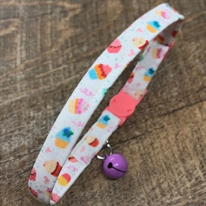 May include: A white cat collar featuring a pattern of colourful cupcakes. It has a pink buckle and a small purple bell. The background is a wooden surface.