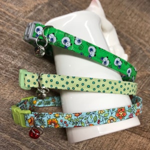 May include: Three colorful cat collars with bell charms. The collars are made of fabric with different patterns: green with white polka dots, green with white cartoon mice, and blue with a floral pattern.