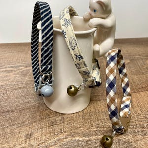 May include: Three patterned cat collars with bells. One is navy blue with white stripes, another has a seashell design, and the third is a brown and navy blue plaid. A white ceramic cat figurine is perched on a white mug.