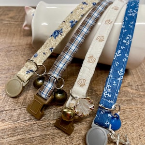 May include: Four patterned cat collars. One is beige with blue floral print, another is brown and blue plaid, a third is beige with seashell print, and the last is blue with white anchors. Each has a bell and a plastic clasp.