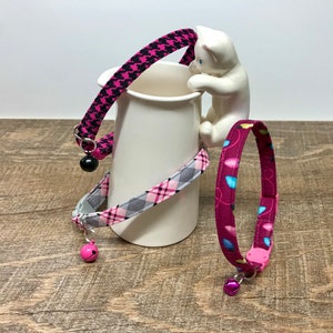 May include: Three cat collars in pink, gray, and purple with various patterns and bell charms. One collar has a black and pink houndstooth pattern, another has a gray and pink plaid, and the third has a purple pattern with blue and teal shapes.