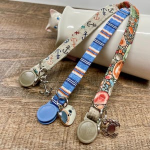 May include: Three patterned pet collars with metal hardware and charms. One collar features blue and white stripes, another has a nautical anchor design, and the third has a floral pattern. Each collar has a different charm, including a crab, anchor, and a cat.