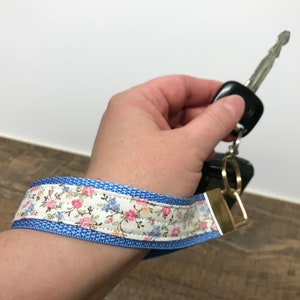 Pode incluir: Um chaveiro de tecido azul e branco com padrão floral e um anel de metal dourado. O chaveiro é feito de tecido e tem uma borda azul.