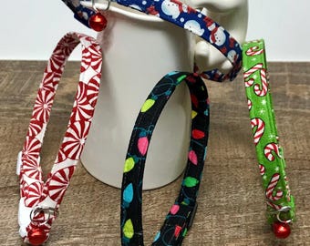 May include: Four festive cat collars with different Christmas patterns: red and white candy canes, green and white candy canes, blue and white snowmen, and black with colorful lights. Each collar has a silver bell attached.