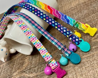 Könnte beinhalten: Vier bunte Katzenhalsbänder mit passenden Glöckchen und Schnallen. Die Halsbänder weisen Muster aus Regenbogenwellen, Herzen, Punkten und Karos auf. Jedes Halsband hat eine unterschiedlich farbige Schnalle und Glocke.