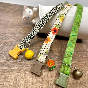May include: Three patterned cat collars with plastic buckles and metal hardware. One collar is black with white polka dots, another is cream with orange flowers, and the third is green with leaf designs. Each has a bell charm.