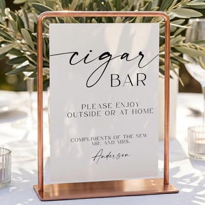 Può includere: Un cartello rettangolare bianco con testo nero che recita cigar BAR, PLEASE ENJOY OUTSIDE OR AT HOME, COMPLIMENTS OF THE NEW MR. AND MRS. Anderson, inserito in un supporto con cornice in metallo color rame su una superficie bianca.
