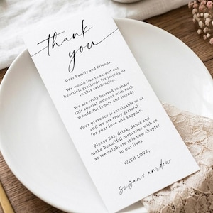 May include: A rectangular white thank you card with black printed text featuring a script header that reads Thank you, followed by a message to family and friends, and signed by Susan and Andrew, placed on a white ceramic plate with gold cutlery.