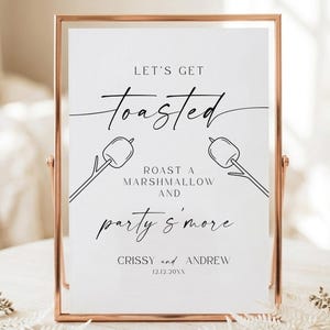 May include: A rectangular sign in a rose gold metal frame featuring the black text LET'S GET Toasted, ROAST A MARSHMALLOW AND party s'more, CRISSY and ANDREW, 12.12.20XX, with line art illustrations of two marshmallows on roasting sticks.
