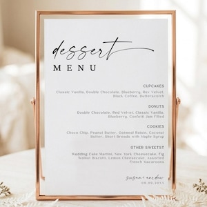 Puede incluir: Una tarjeta de menú de postres blanca en un marco de metal oro rosa. El texto dice dessert MENU, seguido de listas de cupcakes, donas, galletas y otros dulces, terminando con los nombres sugar and dew y la fecha 09.09.20xx.