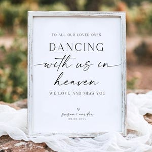 May include: A white rectangular sign in a distressed white wooden frame featuring black text that reads: TO ALL OUR LOVED ONES DANCING with us in heaven WE LOVE AND MISS YOU, followed by the names susan + andrew and the date 09.09.20XX.