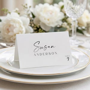Könnte beinhalten: Eine weiße, gefaltete Tischkarte mit dem schwarz gedruckten Namen Susan Anderson und dem Text Table 7. Die Karte steht auf einem weißen Speiseteller mit Goldrand und bietet ein klares, schlichtes Design für die Sitzordnung bei Veranstaltungen.